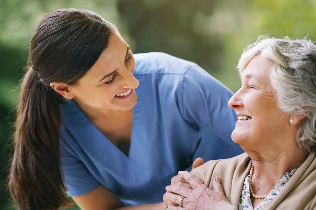 A Nurse Is Smiling As She Helps An Older Woman.