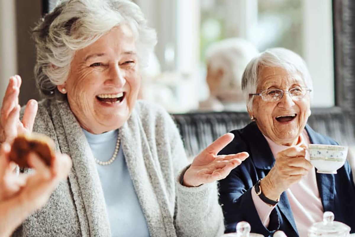 Three Older Women Laughing And Eating At A Table.